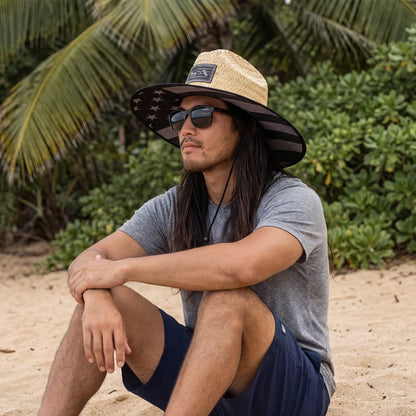 Man sitting on a beach wearing a wide-brimmed hat and sunglasses with palm trees in the background.