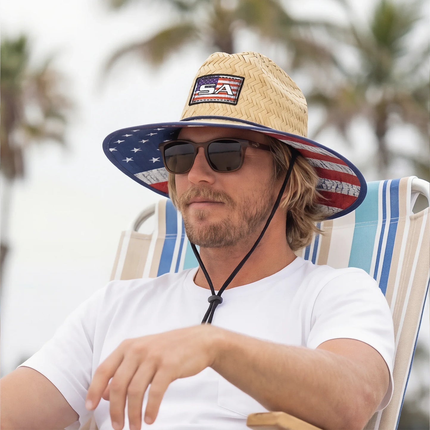 Man wearing a straw hat with an American flag design, sunglasses, and a white t-shirt, sitting on a striped chair.