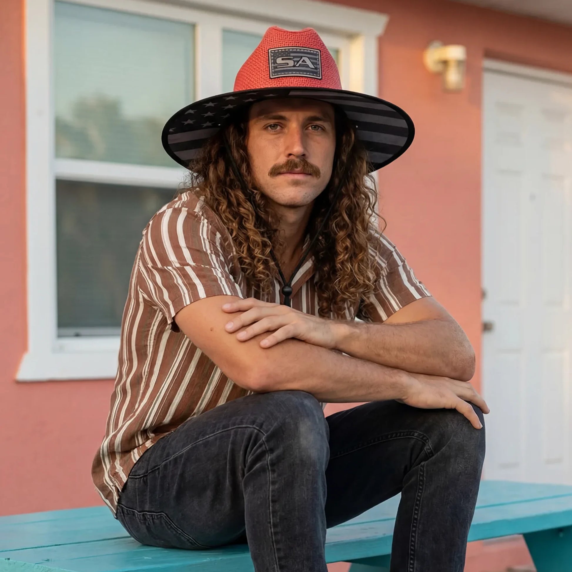 Man wearing a red and black hat sitting on a teal bench in front of a pink building.