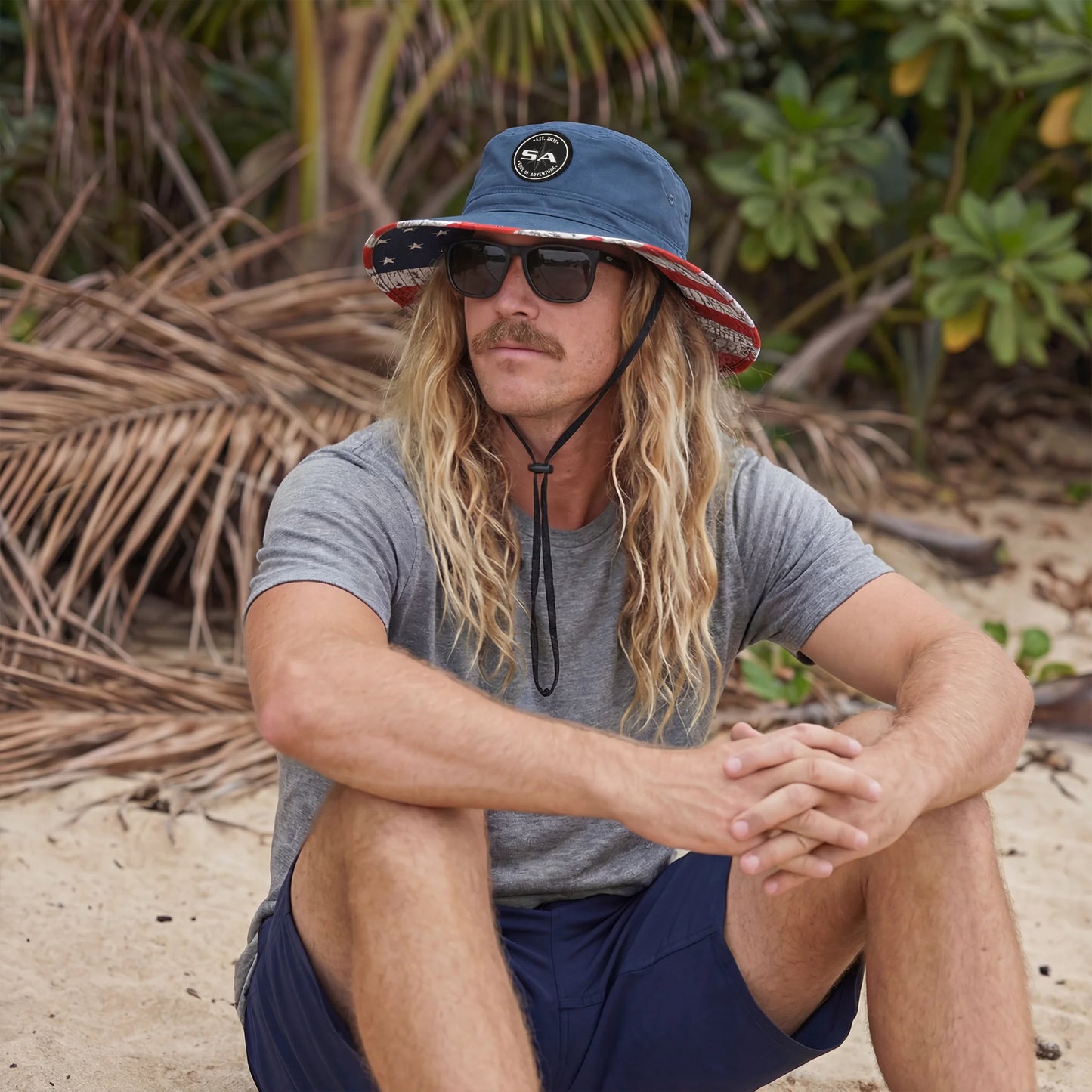 Man sitting on a beach wearing sunglasses, a gray t-shirt, and a blue bucket hat.