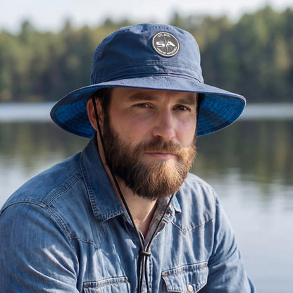 Man wearing a blue bucket hat with a logo, sitting by a lake with trees in the background.