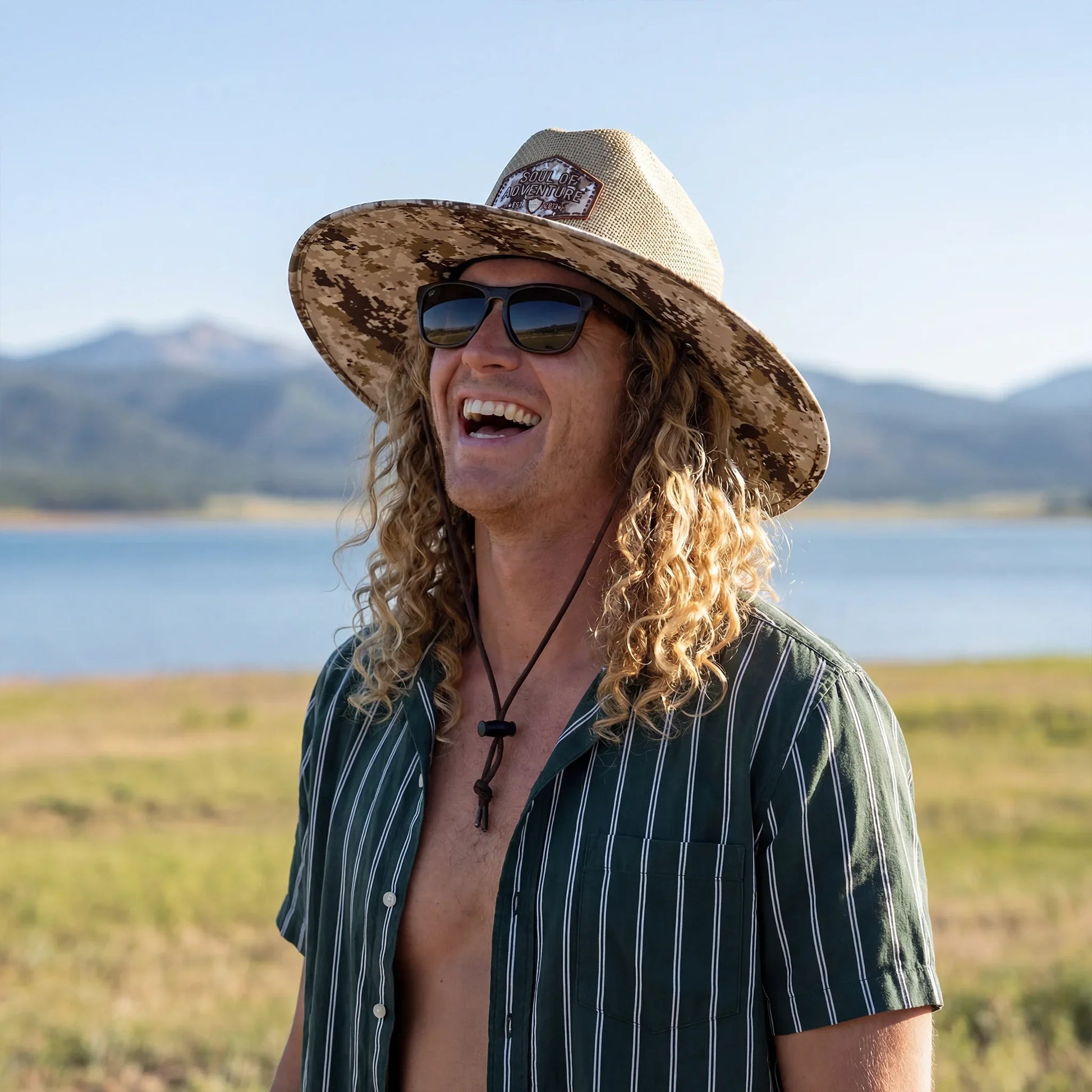 Man wearing a wide-brimmed hat and sunglasses, standing in a scenic outdoor setting with mountains and water.